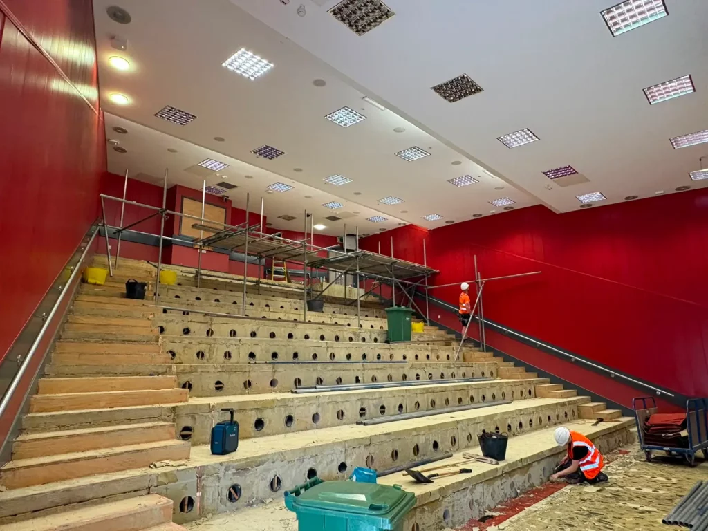 The initial framework of an internal birdcage scaffold being erected over steep tiered seating inside a university lecture theatre.